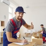 Happy smiling delivery company worker guarantees best quality service. Portrait of cheerful handsome bearded young man in uniform workwear filling in order papers, looking at camera, showing thumbs up